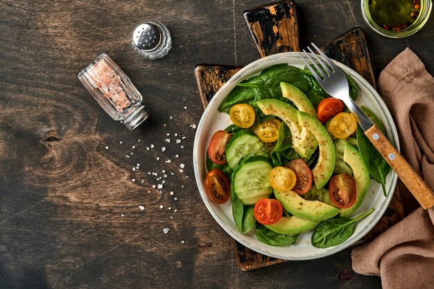 Avocado, rote, gelbe, schwarze Kirschtomate, Spinat und frischer Salat der Gurke mit Gewürzpfeffer und Olivenöl im alten Holztischhintergrund der grauen Schüssel Gesundes Lebensmittelkonzept. Ansicht von oben.