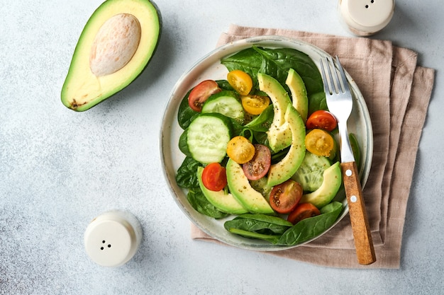 Avocado, rote, gelbe, schwarze Kirschtomate, Spinat und Gurke frischer Salat mit Gewürzpfeffer und Olivenöl in grauer Schüssel auf grauem Schiefer-, Stein- oder Betonhintergrund. Gesundes Lebensmittelkonzept. Ansicht von oben.