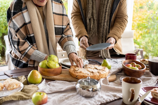 Avós contemporâneos em trajes casuais aconchegantes ao lado da mesa enquanto o servem antes do jantar, mulher cortando pão e homem ajudando-a