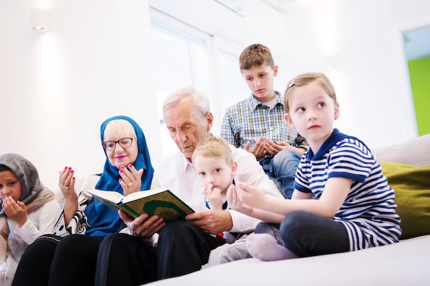 avós da família muçulmana moderna com netos lendo o Alcorão e orando juntos no sofá antes do jantar iftar durante uma festa do ramadã em casa