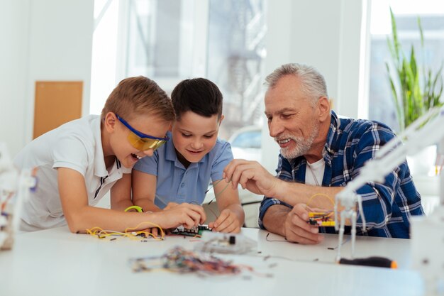 Ayuda y apoyo. Profesor alegre positivo sentado junto con sus alumnos mientras les ayuda a construir un robot