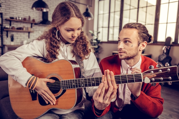 Ayudando a la hermana. Hermano mayor cariñoso barbudo ayudando a su linda hermana adolescente tocando la guitarra