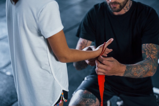 Ayudando a llevar los vendajes. Entrenador de boxeo tatuado joven enseña a niña linda en el gimnasio.