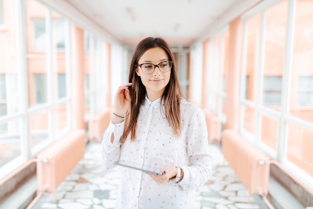 Ayudante de sexo femenino sonriente de los jóvenes con las lentes y la tableta de la tenencia del pelo marrón y el pelo conmovedor mientras que se coloca en el pasillo.