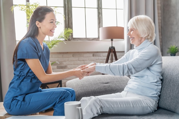 Ayudar a las personas mayores Cuidadora sosteniendo las manos de una anciana en el interior