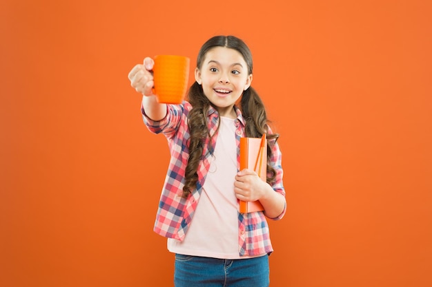 Ayúdate a ti mismo Linda colegiala disfrutando de las vacaciones escolares sobre fondo naranja Niña pequeña tomando un descanso para el té Niño pequeño sosteniendo un libro y una taza en el descanso para comer Es tiempo de descanso