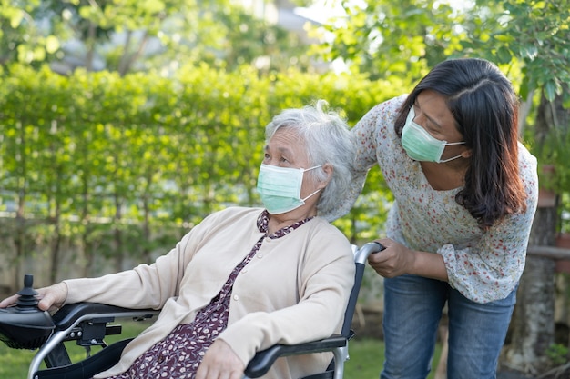 Ayude a una anciana asiática mayor o anciana en silla de ruedas eléctrica y con una mascarilla para proteger la infección de seguridad Covid-19 Coronavirus en el parque.