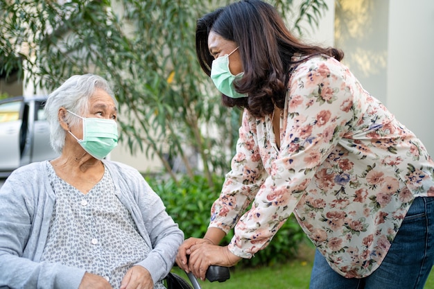 Ayude a una anciana asiática mayor o anciana en silla de ruedas y con una máscara facial para proteger la infección de seguridad Covid-19 Coronavirus en el parque.