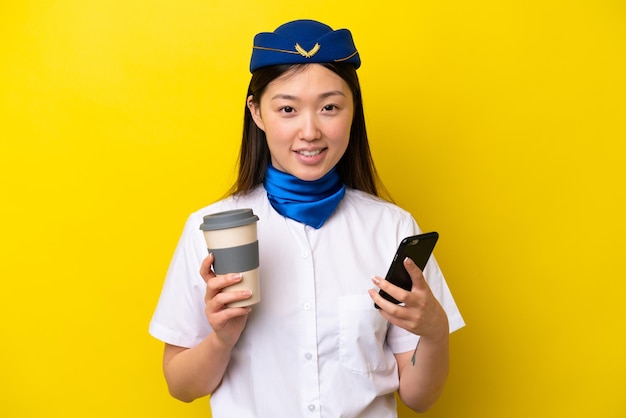 Azafata de avión china aislada de fondo amarillo sosteniendo café para llevar y un móvil