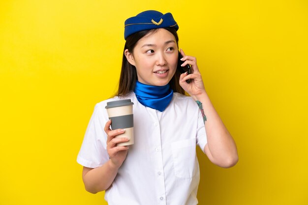 Azafata de avión china aislada de fondo amarillo sosteniendo café para llevar y un móvil