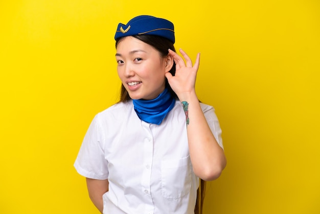 Azafata china de avión aislada de fondo amarillo escuchando algo poniendo la mano en la oreja