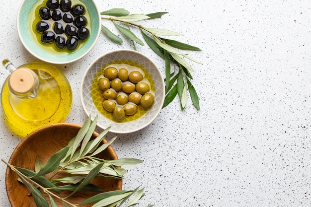 Azeitonas verdes e pretas com azeite em uma garrafa de vidro, raminhos de oliveira e pão de ciabatta fresco cortado em uma tábua de madeira. Fundo branco rústico, comida mediterrânea saudável, espaço para texto