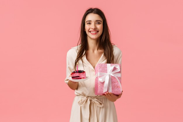 B-day girl se siente feliz y optimista, disfruta celebrando. Atractiva mujer caucásica recibe regalos, sosteniendo un delicioso pastel, envuelto presente, sonriendo alegremente, de pie en la pared rosa