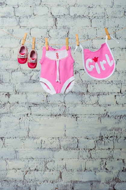 Foto babero de cuerpo rosa para niños y zapatos rojos secos sobre una cuerda contra una pared de ladrillo blanco.