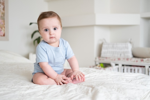 Baby 6 Monate alt im blauen Bodysuit lächelnd und sitzend auf weißem Bett zu Hause.