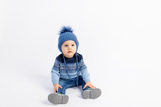 Baby 8 Monate alter Junge sitzt auf einer weißen isolierten Wand in warmer Winterkleidung und einem Hut, Kindermode, Werbung für Kinderkleidung,