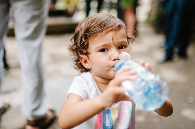 Baby Baby mit braunem Haar trinkt Wasser im Park mit Plastikflasche Kleines Mädchen, das im Freien auf der Straße geht