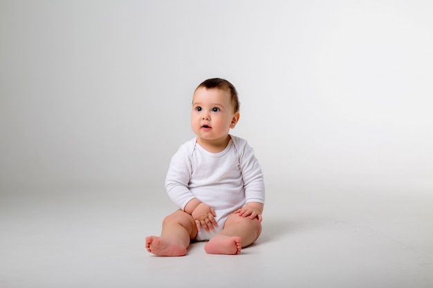 Foto baby boy 9 meses en un mono blanco sentado en una pared blanca