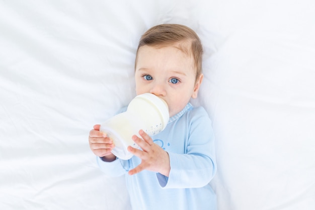 Baby Boy come leche de un biberón en la cama antes de irse a la cama con un traje azul, concepto de comida para bebés