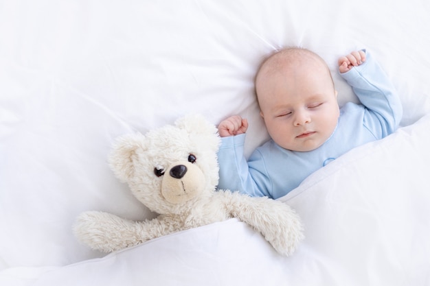 Baby Boy duerme en la cama acostado boca arriba con un oso de peluche en pijama azul