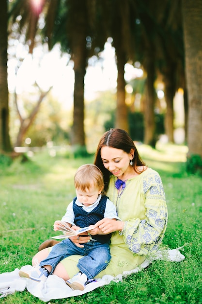 Baby Boy está sentado en el regazo de su mamá y leyendo un libro en un picnic en el parque