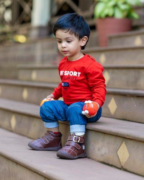 Baby Boy jugando con juguetes sentado en las escaleras vistiendo camiseta roja y jeans azules