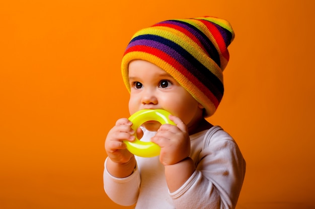 Baby Boy en un mono blanco y sombrero multicolor sosteniendo un juguete sentado en una pared amarilla