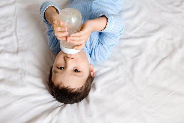 Baby Boy Morena se encuentra en la cama en pijama azul y bebe leche de una botella. Comida para bebé