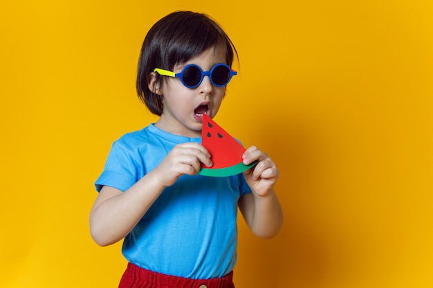 Baby Boy en pantalones rojos come una sandía de papel en una pared amarilla