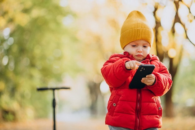 Baby Boy en scooter sosteniendo teléfono móvil en el parque otoñal