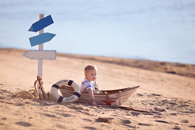 Baby Boy sentado en un barco, vestido como un marinero, en una playa de arena con conchas junto al mar