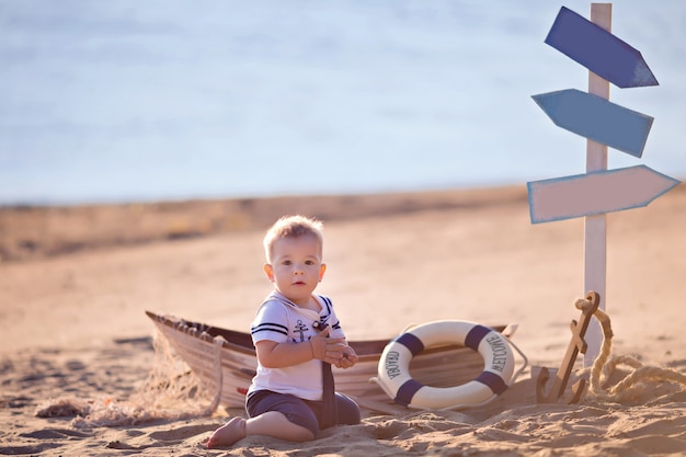 Baby Boy sentado en un barco, vestido como un marinero, en una playa de arena con conchas junto al mar