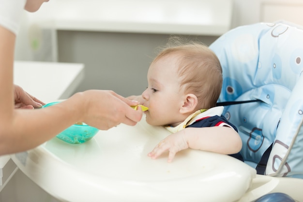 Baby Boy sentado en la trona en la cocina y comiendo de una cuchara