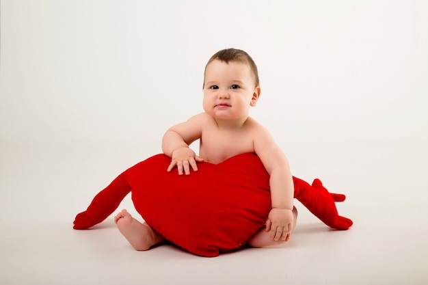 Baby Boy sonriendo y sosteniendo una almohada roja en forma de corazón, sentado en una pared blanca