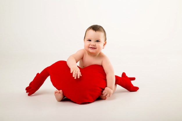 Baby Boy sonriendo y sosteniendo una almohada roja en forma de corazón, sentado en una pared blanca