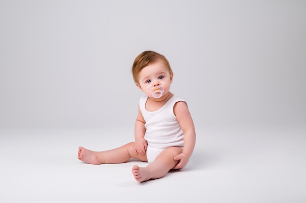 Baby Boy en un traje blanco con un chupete en la boca está sentado sobre un fondo blanco.