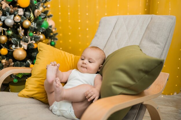 Baby Boy en un traje blanco sentado en una mecedora con árbol de Navidad