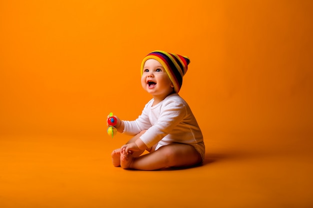 Foto baby boy en un traje blanco y sombrero multicolor con un juguete, sentado en una pared de color naranja