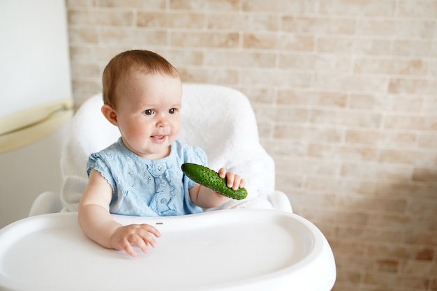 Baby, das Gemüse isst. grüne Gurke in der Hand des kleinen Mädchens in der sonnigen Küche.