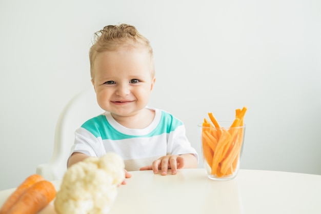 Baby, das in einem Kinderstuhl sitzt und Karottenscheiben auf weißem Hintergrund isst