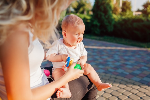 Baby, das mit den Spielwaren sitzen auf den Knien ihrer Mutter im Sommerpark spielt.