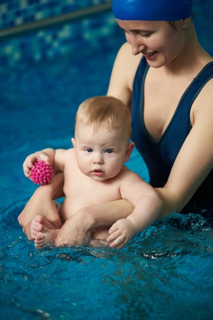 Baby hält Spielzeugbälle und sitzt während Wellnessbehandlungen in den Händen seiner Mutter im klaren blauen Wasser des Swimmingpools