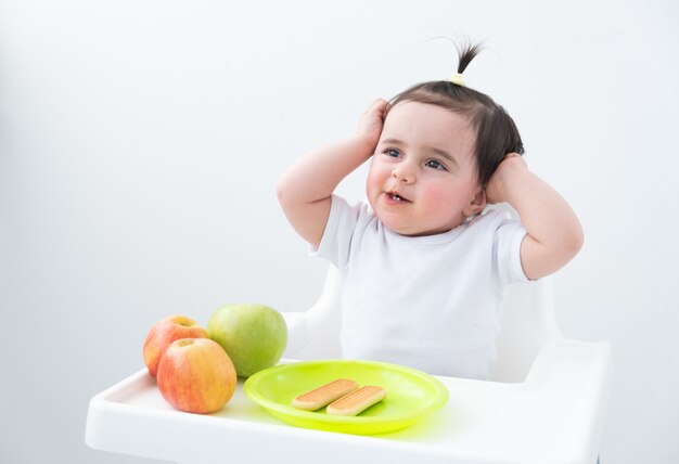 Baby im Babystuhl, der Äpfel und Kekse isst. Baby erste feste Nahrung.