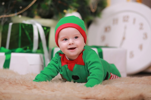 Foto baby im rotgrünen elfenkostüm, das unter weihnachtsbaum und geschenkboxen sitzt.