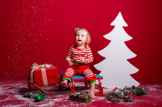 Baby im Weihnachtspyjama und in der Weihnachtsmütze fängt den Schnee, der auf einem Schlitten mit Geschenkbox und großem weißem Weihnachtsbaum auf rotem Hintergrund sitzt