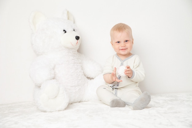 Baby im weißen Anzug sitzt mit großem weißen Teddybären und schaut in die Kamera auf weißem Hintergrund.