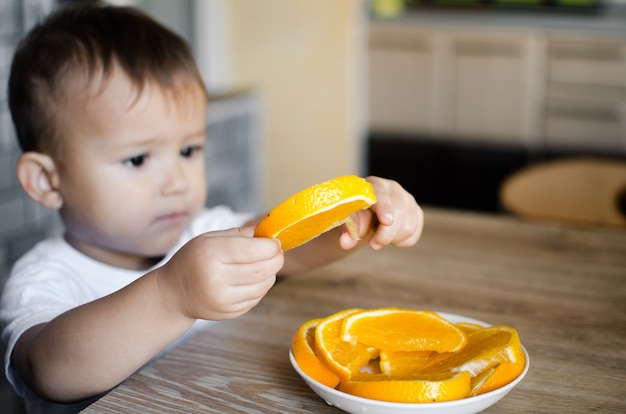 Baby in der Küche in einem weißen T-Shirt nimmt eine Orangenscheibe