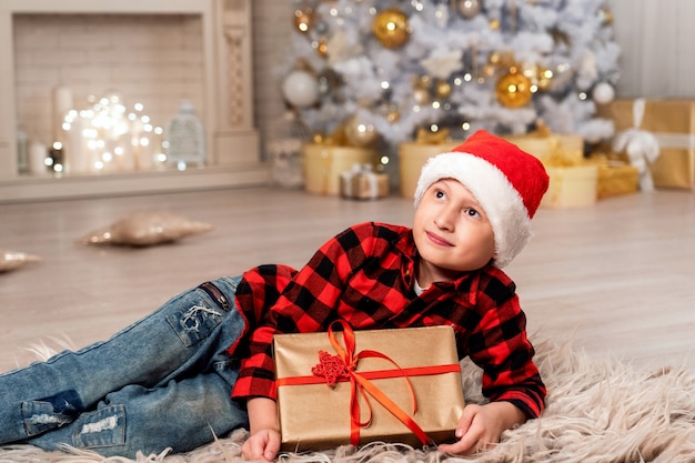 Baby in der Weihnachtsmütze, die breit lächelnd schaut. Das Zimmer ist mit Weihnachtslichtern dekoriert. eleganter schneebedeckter Weihnachtsbaum im Fokus. Familienurlaub Weihnachten gefällt dem Kind