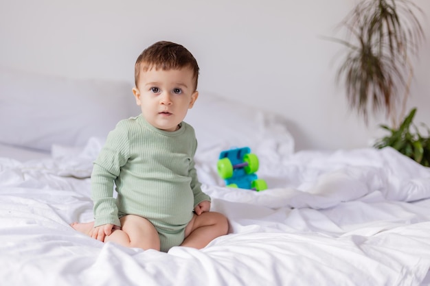 Baby in einem grünen Baumwollbody sitzt auf einem weißen Bett mit einem farbigen Spielzeugautokind spielt