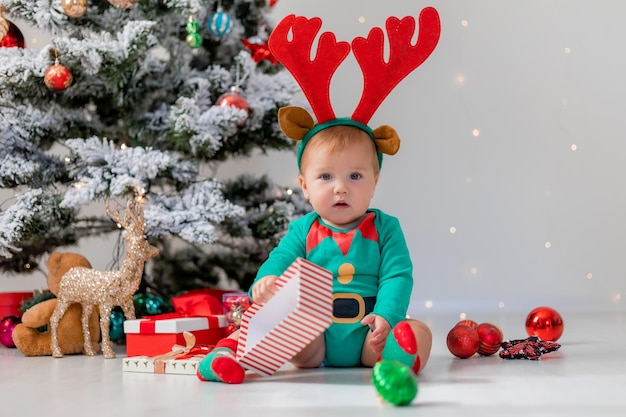 Baby in einem grünen Body mit roten Hirschhörnern auf dem Kopf spielt mit Geschenkboxen in der Nähe des Weihnachtsbaums. Kind in einem Weihnachtszwergkostüm. Winter-Neujahrskonzept. Urlaubsware. Platz für Text